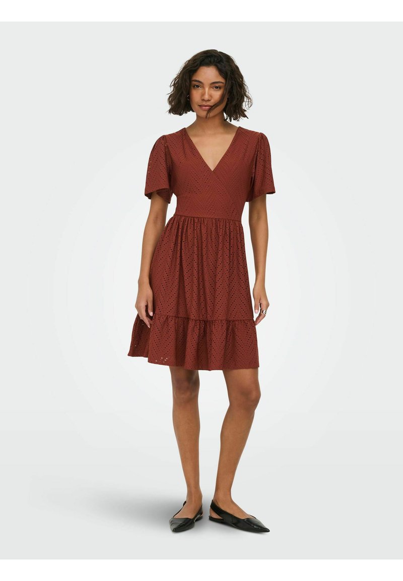 Femme debout portant une robe à manches courtes, longueur genou, de couleur rouille avec des ajours, col en V, et des ballerines noires pointues.