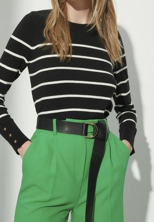 Femme portant une chemise à manches longues rayée noir et blanc rentrée dans un pantalon taille haute vert vif, avec une ceinture noire et les mains dans les poches.