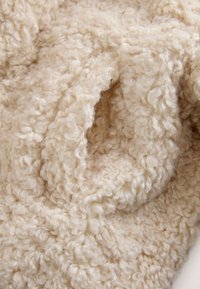 Bézs színű, texturált anyag, amely göndör hurkokat és puha, bolyhos felületet mutat. Az anyag melegnek és kényelmesnek tűnik, puha textúrával.