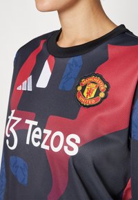 Svart langermet genser med et dristig geometrisk design i rødt og blått. Den viser Manchester United-logoen og hvit "Tezos"-tekst.