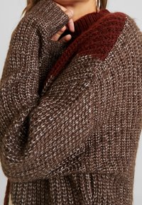 Paksu neulepusero ruskeana, jossa on sekoitettu pinta, burgundinpunainen olkapäällywoodistus ja pitkät, ribatut hihat.