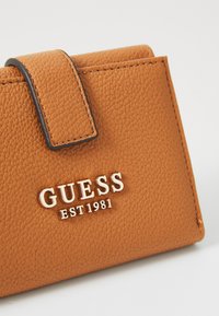 Narančasti novčanik teksturiranog dizajna s magnetskim zatvaranjem, s uzdignutim zlatnim logotipom "GUESS EST 1981" sprijeda. Glatka, elegantna završna obrada.