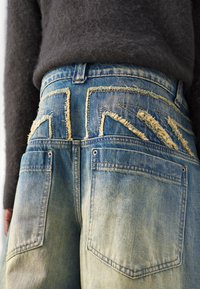 Jeans in denim con un colore blu sbiadito, dettagli strappati e modelli di cuciture unici sulle tasche posteriori.