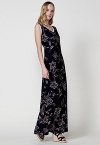 Robe maxi en tissu bleu marine ornée de broderies florales argentées et rouges, avec un décolleté en V et un design sans manches, accompagnée de sandales compensées.