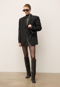 Woman wearing oversized black blazer, gray button-up shirt, black shorts, sheer tights, knee-high black boots, rectangular sunglasses, and a small black handbag.