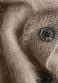 Beiges gestricktes Pulloverwarenstoff mit strukturierter Oberfläche, versehen mit einem großen schwarzen Knopf mit vier Löchern und weichen, flauschigen Fasern.