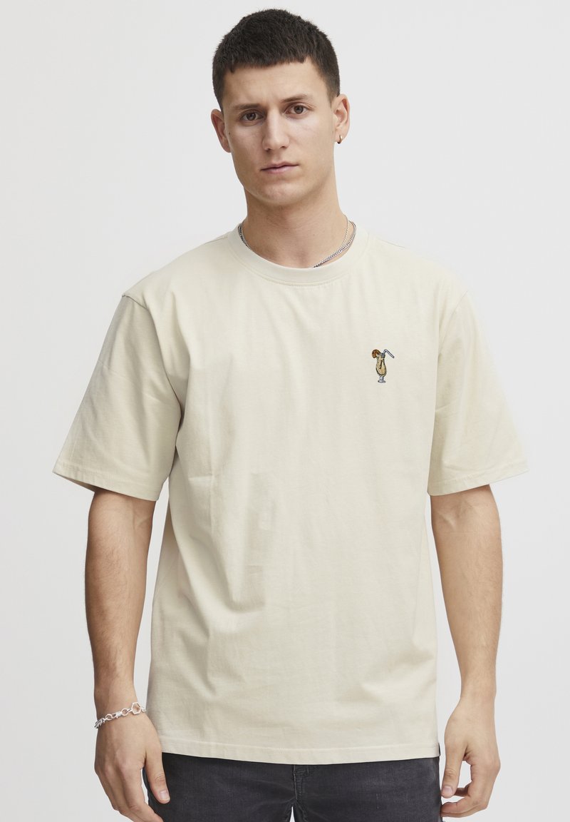 Beige cotton t-shirt with short sleeves, featuring a small embroidered design on the chest. Simple and relaxed fit.