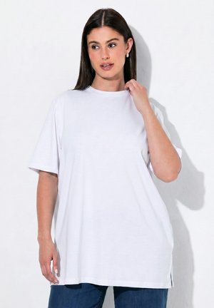 Frau mit langen dunklen Haaren, die ein lockeres weißes T-Shirt und blaue Jeans trägt, steht vor einem weißen Hintergrund.
