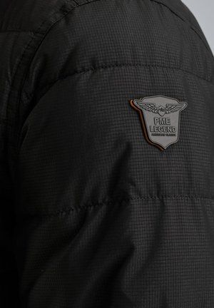 Doudoune noire avec un tissu texturé, un design matelassé et un logo PME Legend en forme de bouclier sur l'épaule gauche.