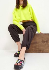 Neonowo zielona krótka bluza z kapturem, czarne zwężane spodnie z elastycznymi ściągaczami oraz sandały na platformie z kolorowymi wzorami graficznymi po bokach.