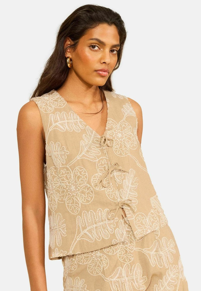 Donna che indossa un top beige senza maniche e pantaloncini con ricamo floreale bianco e chiusure frontali annodate, in posa su sfondo semplice.