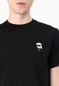 Černé bavlněné tričko se cartoon broží znázorňující postavu v obleku s bílými vlasy a slunečními brýlemi, umístěnou v levé části hrudníku.
