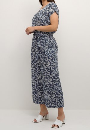 Femme portant une combinaison bleue à motif floral avec des manches courtes et des sandales blanches à talons blocs ouvertes aux orteils, debout de profil.