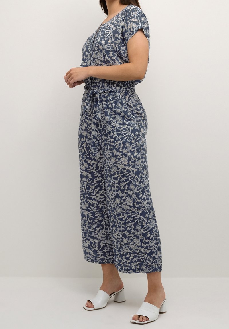 Femme portant une combinaison bleue à motif floral avec des manches courtes et des sandales blanches à talons blocs ouvertes aux orteils, debout de profil.