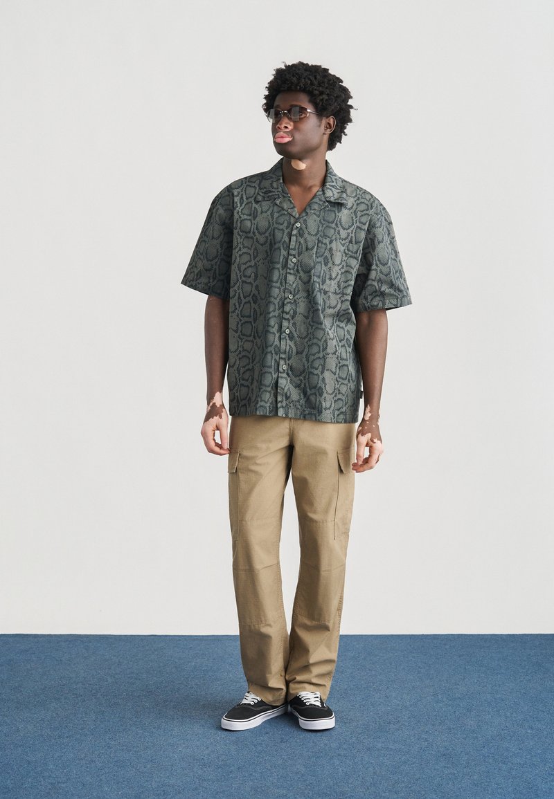 Camicia verde scuro a maniche corte con bottoni e stampa testurizzata a motivo serpente, abbinata a pantaloni cargo beige chiaro e sneakers nere.