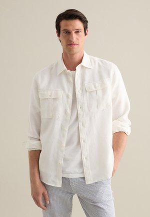 Camisa de lino blanca con dos bolsillos frontales, mangas enrolladas y diseño de botón, combinada con pantalones a rayas.