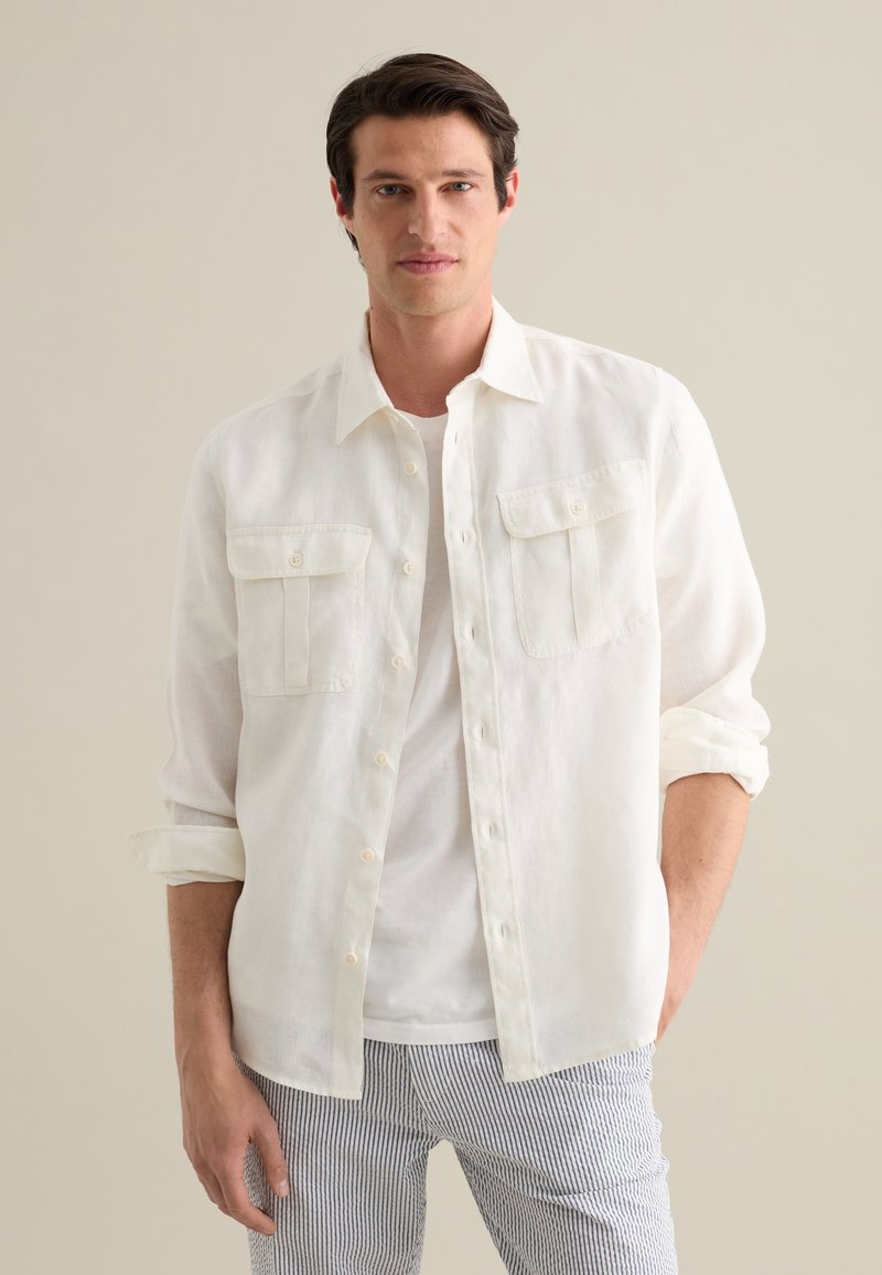 Camicia bianca in lino con due tasche frontali, maniche arrotolate e design con bottoni, abbinata a pantaloni a righe.