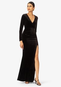 Schwarzes Samtkleid mit langen Ärmeln, tiefem V-Ausschnitt und seitlichem Schlitz, mit gerafften Details an der Taille, ergänzt durch Riemchenhighheels.