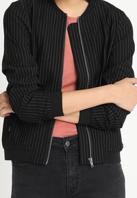 Czarna bomberka w paski z zamek z przodu, ściągaczami przy mankietach i dolnym wykończeniu. Nosi się ją na czerwonej koszulce i ciemnych dżinsach, o gładkiej fakturze.