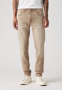 Jeans de denim bege claro com um corte descontraído, apresentando cinco bolsos e fecho de botão. Combinados com sapatilhas brancas.