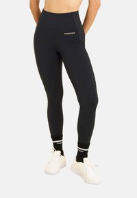 Leggings neri a vita alta realizzati in materiale elasticizzato. Presentano un marchio discreto e una texture liscia. Abbinati a sneakers bianche e calze nere.