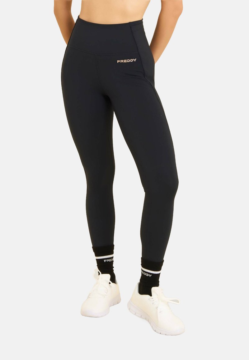 Leggings negros de cintura alta hechos de material elástico. Presentan una marca discreta y una textura suave. Combinados con zapatillas blancas y calcetines negros.