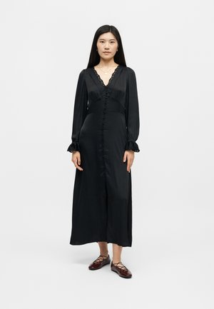 Γυναίκα στέκεται μπροστά σε λευκό φόντο, φοράει μακρύ μαύρο φόρεμα με κουμπιά και δαντελωτό τελείωμα, καθώς και μπορντό πλατφόρμες με κορδόνια.