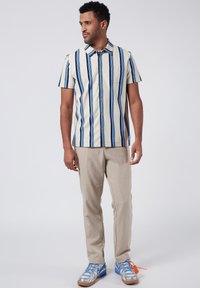 Camicia a maniche corte con righe verticali blu e bianche, chiusura frontale a bottoni, design con colletto, abbinata a pantaloni beige e sneaker blu.