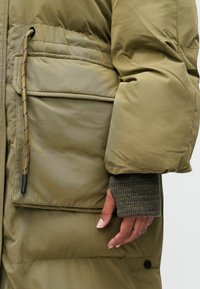 Maslena zeleni puffer kaput s velikim džepom, rebrastim rukavima u sivoj boji i vezicom s crnim detaljima. Glatka, k Dachbarren strukturirana tekstura.