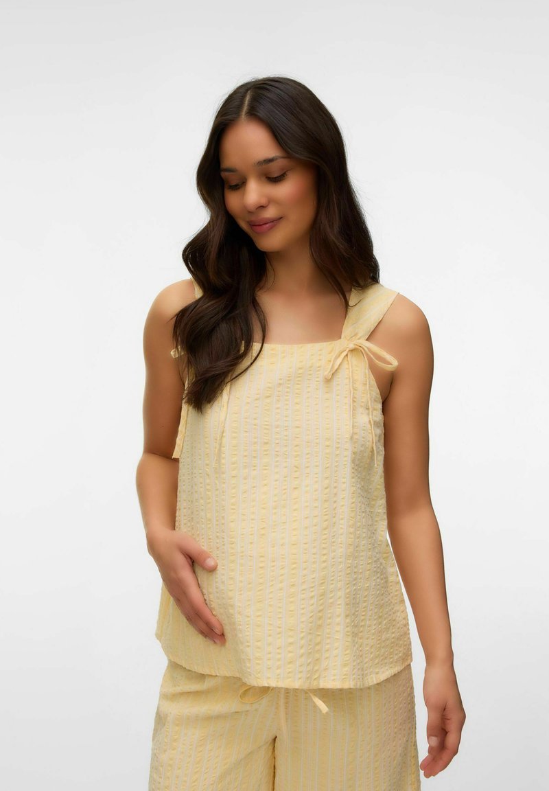 Femme aux longs cheveux foncés portant un haut sans manches jaune et un pantalon, posant délicatement sa main sur son ventre, sur un fond uni.