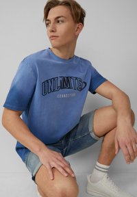Blauwe katoenen t-shirt met korte mouwen, voorzien van een grafische print met de tekst "ONBEPERKTE VERBINDING" in zwart, gecombineerd met denim short.
