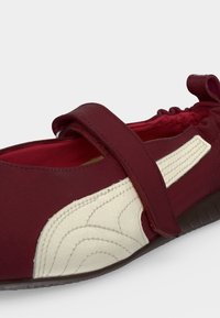 Σκούρο κόκκινο παπούτσι τύπου slip-on με λευκό ραμμένο σχέδιο, ρυθμιζόμενο λουράκι στο πάνω μέρος και ελαστική πίσω πλευρά για ασφαλή εφαρμογή.