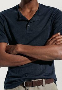 Ciemnoniebieska koszulka polo z małym kołnierzykiem i dwoma guzikami, zestawiona z brązowym skórzanym paskiem. Materiał wydaje się gładki, bez widocznych wzorów.