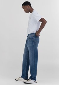 Jeans bleus à jambe droite avec un lavage moyen, design à cinq poches et coutures discrètes. Portés avec un t-shirt blanc simple et des baskets.