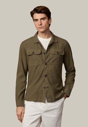 Olivgrünes, leichtes Hemd mit langen Ärmeln, zwei Brusttaschen und schwarzen Knöpfen. Getragen über einem weißen T-Shirt mit Rundhalsausschnitt.