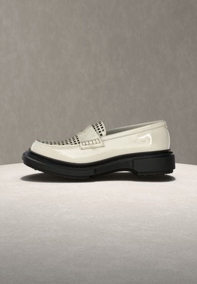 Λευκό δερμάτινο μοκασίνι από λουστρίνι με μαύρη, χοντρή σόλα και πλεκτή λεπτομέρεια στην κορυφή, εμφανιζόμενο σε πλαϊνή όψη σε ουδέτερη επιφάνεια.