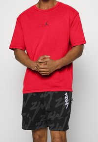 Punainen puuvillapaita, jossa on pyöreä kaula-aukko ja lyhyet hihat, varustettuna mustalla Jumpman-logolla. Yhdistetty mustiin, kuviollisiin shortseihin, joissa on harmaita yksityiskohtia.