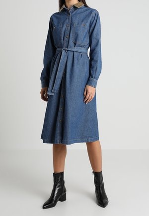 Kobieta w długiej do kolan sukience koszulowej z denimu z długimi rękawami, związaną w talii paskiem, z przodu zapinanej na guziki, w czarnych botkach za kostkę.