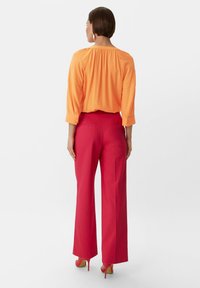 Orange, lockere Bluse mit gerafftem Ausschnitt, kombiniert mit hochgeschnittenen, weiten roten Hosen. Glatte Textur und klare Linien. Rote Absätze setzen Akzente.