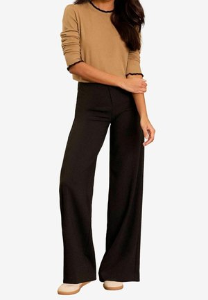 Femme portant un pull beige à bordure noire et un pantalon noir large taille haute, debout, les bras croisés.