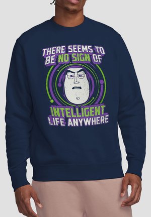 Mann trägt einen marineblauen Pullover mit einem Cartoon-Gesicht und dem Text: "Es scheint nirgendwo Anzeichen intelligenten Lebens zu geben" in Grün und Lila.