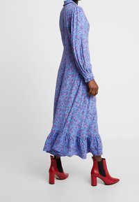 Dlouhé modré květinové šaty s růžovými akcenty, nařasenými rukávy a volánovým lemem, spárované s červenými kozačkami do kotníků s černým elastickým panelem.