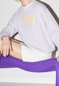 Jasnofioletowy sweatshirt z logo "LEVI'S" w kolorze żółtym, w połączeniu z białymi szortami i długimi fioletowymi skarpetkami. Gładkie tekstury na całej powierzchni.