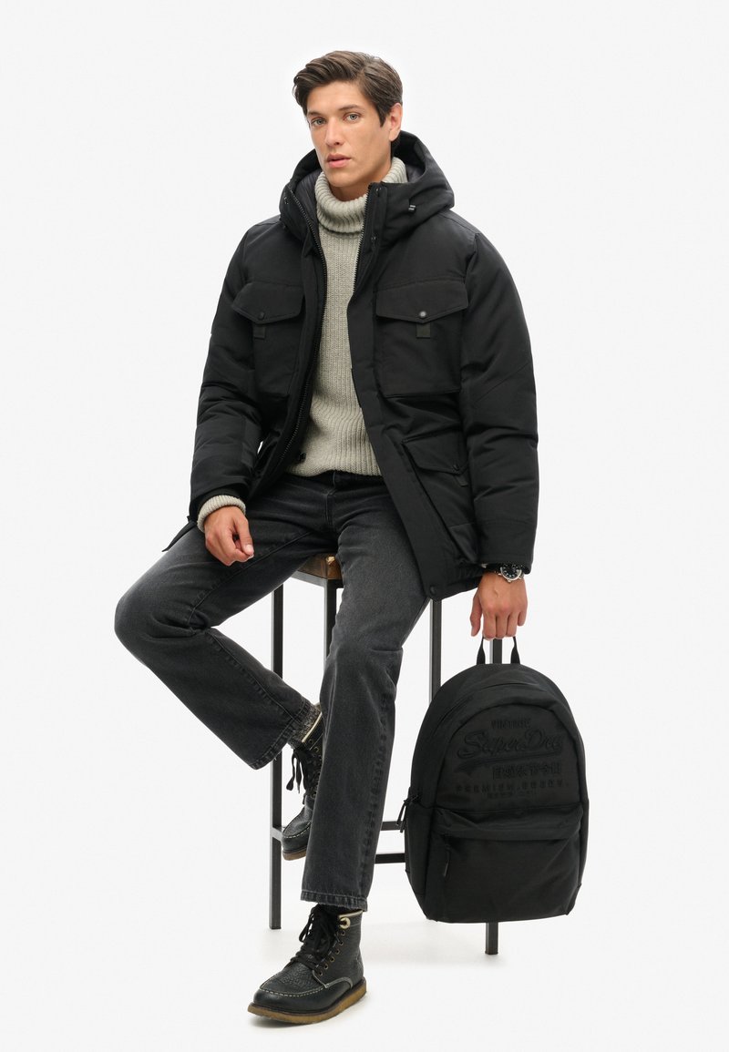 Veste rembourrée noire avec capuche, pull en knit gris en dessous, jean noir et bottes noires à lacets. Un sac à dos noir repose à ses côtés.