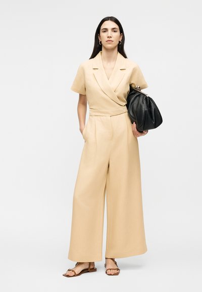 Γυναίκα που στέκεται με τα χέρια στις τσέπες, φοράει μπεζ ολόσωμη φόρμα με φαρδιά πόδια, καφέ σανδάλια, χρυσούς κρίκους για σκουλαρίκια και κρατάει μεγάλη μαύρη τσάντα κάτω από το χέρι.