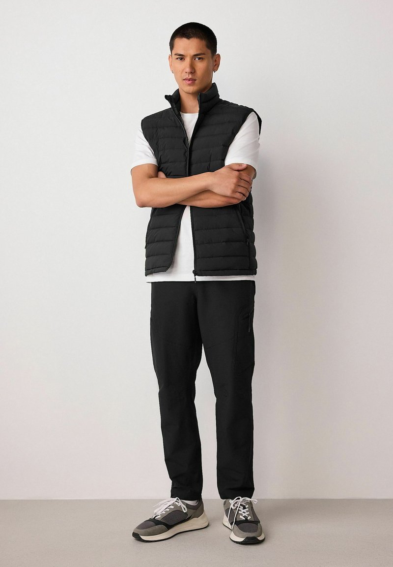 Black puffer vest with a high collar, worn over a white t-shirt. Paired with black pants and gray athletic shoes with white laces.