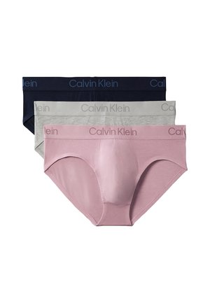  umięśniony mężczyzna stojący boso i zwrócony przodem, ubrany w beżowe majtki Calvin Klein na jednolitym szarym tle.