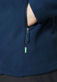 Tamsiai mėlynas švarkas iš vilnos, su užtrauktuku ir kišenėle, pasižymintis tekstūruota paviršiumi bei mažu žaliu žymekliu. Užtrauktukas ir siūlės yra matomos.