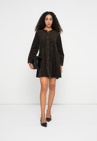 Abito camicia in corduroy marrone con bottoni frontali, maniche lunghe e gonna svasata. Abbinato a una piccola borsa a tracolla nera e ballerine nere.