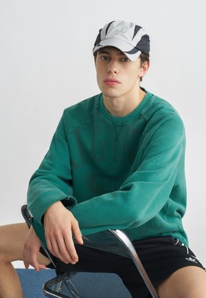 Mladý človek sediaci na priehľadnej stoličke, oblečený v zelenom pulóvri, čiernych šortkách a bielo-čiernej šiltovke s neutrálno výrazom tváre.